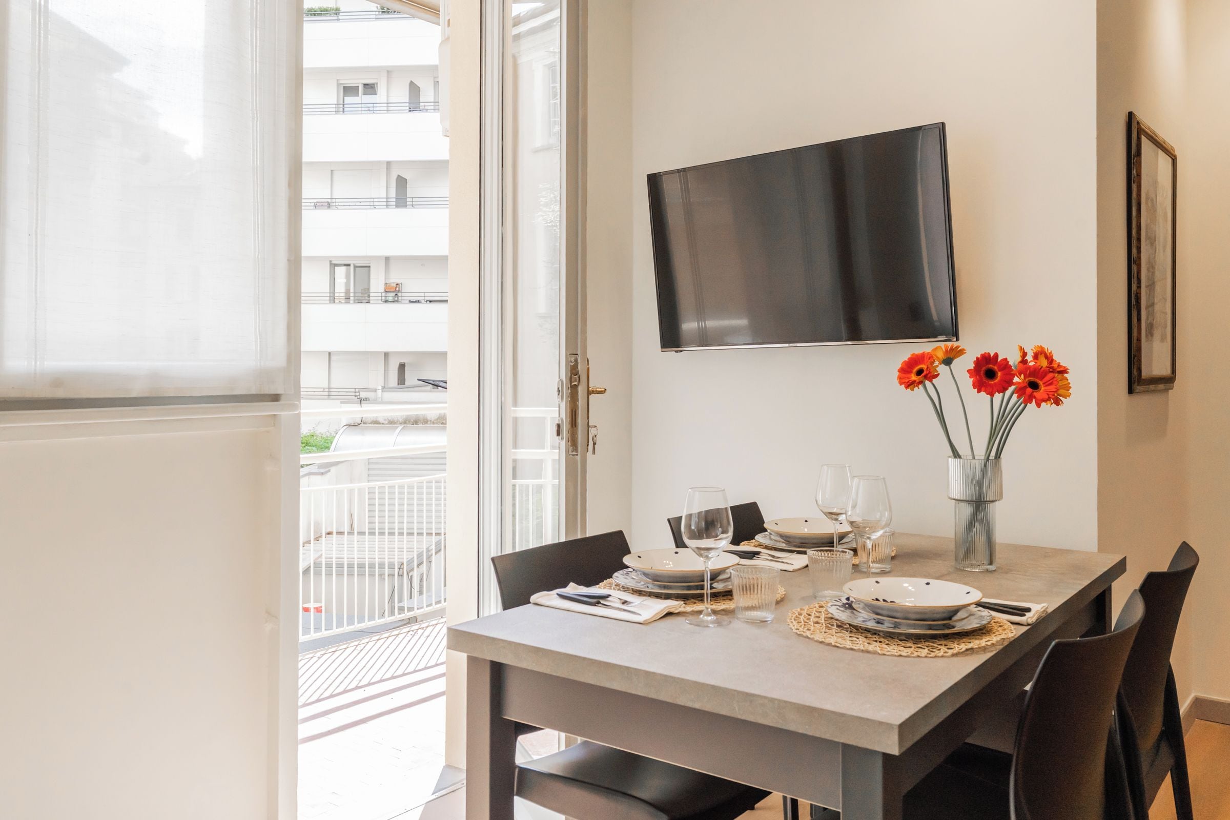 A dining area with a table set for two, a flat-screen TV mounted on the wall, and a vase with flowers on the table.