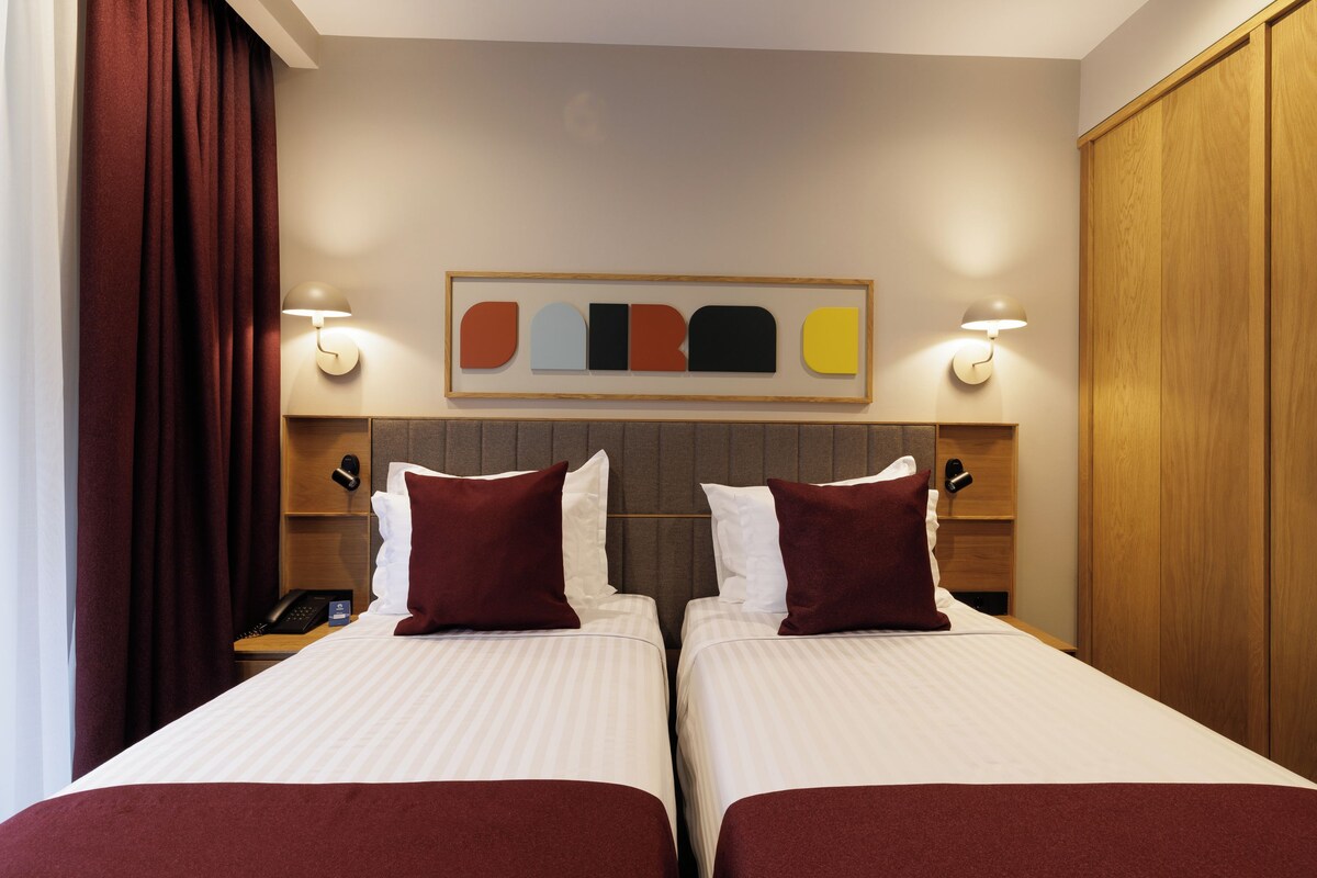 Habitación de hotel con dos camas, cabecera con diseño geométrico, lámparas de noche, teléfono y una obra de arte colgada en la pared.