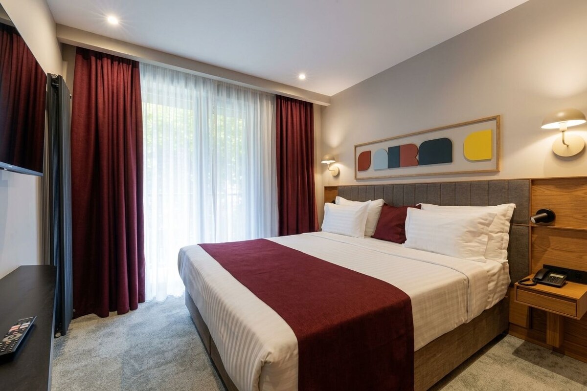 Habitación de hotel con una cama grande, una mesita de noche con lámpara, un televisor y un ventanal con cortinas.