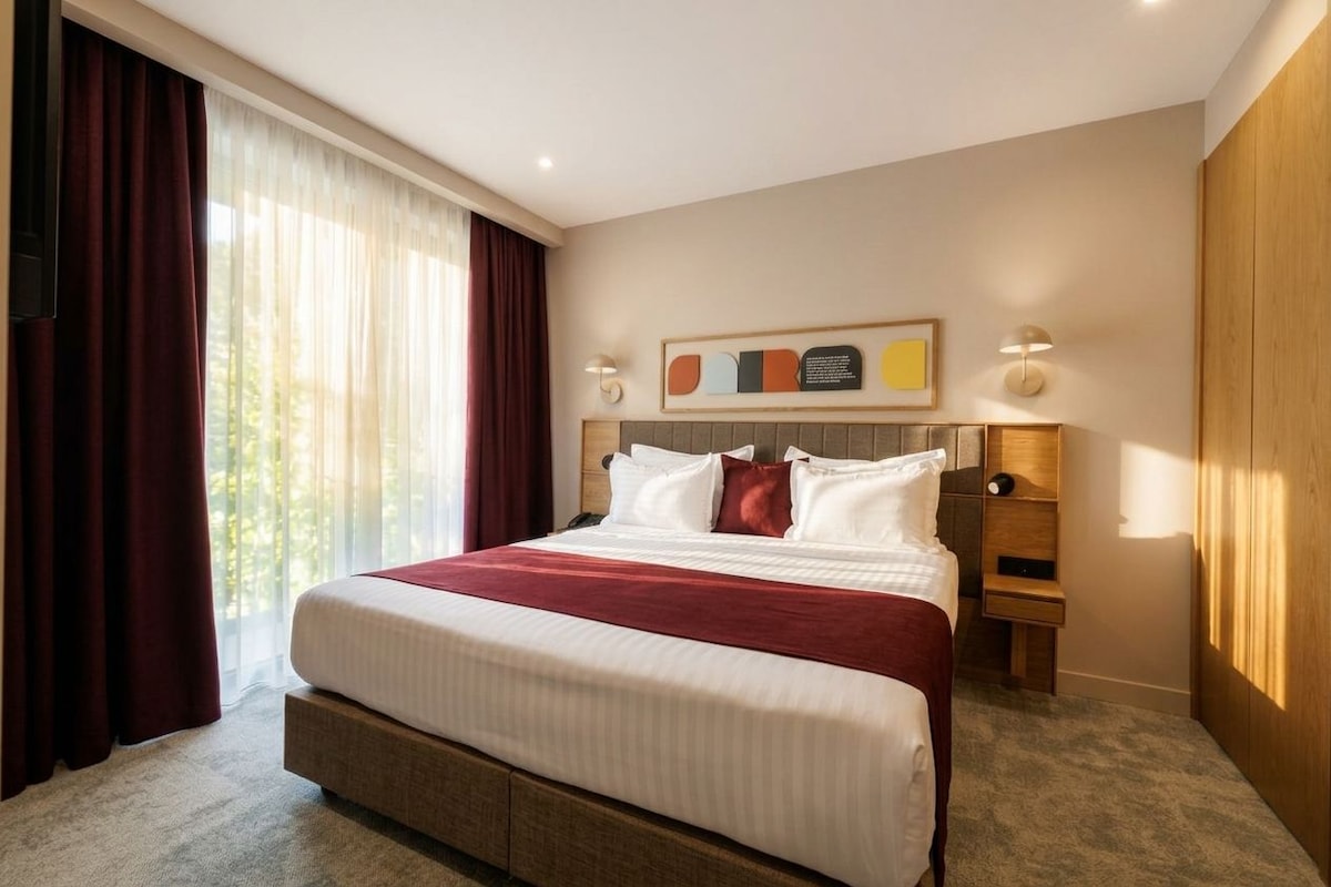 Habitación de hotel con una cama grande, una mesita de noche, un cuadro colgado en la pared y una ventana con cortinas.