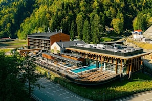 Un hotel con zona de piscina, un edificio grande de fachada de ladrillo y un cerro cubierto de bosque al fondo.