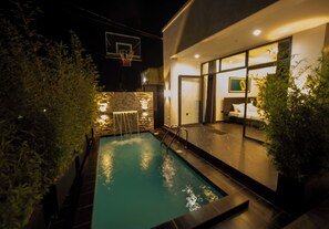 A swimming pool with a waterfall feature, a basketball hoop, and a well-lit interior space with a bed and a framed picture. - Adorable 4-bedroom house in Kigali perfect for family stays (Kigali)