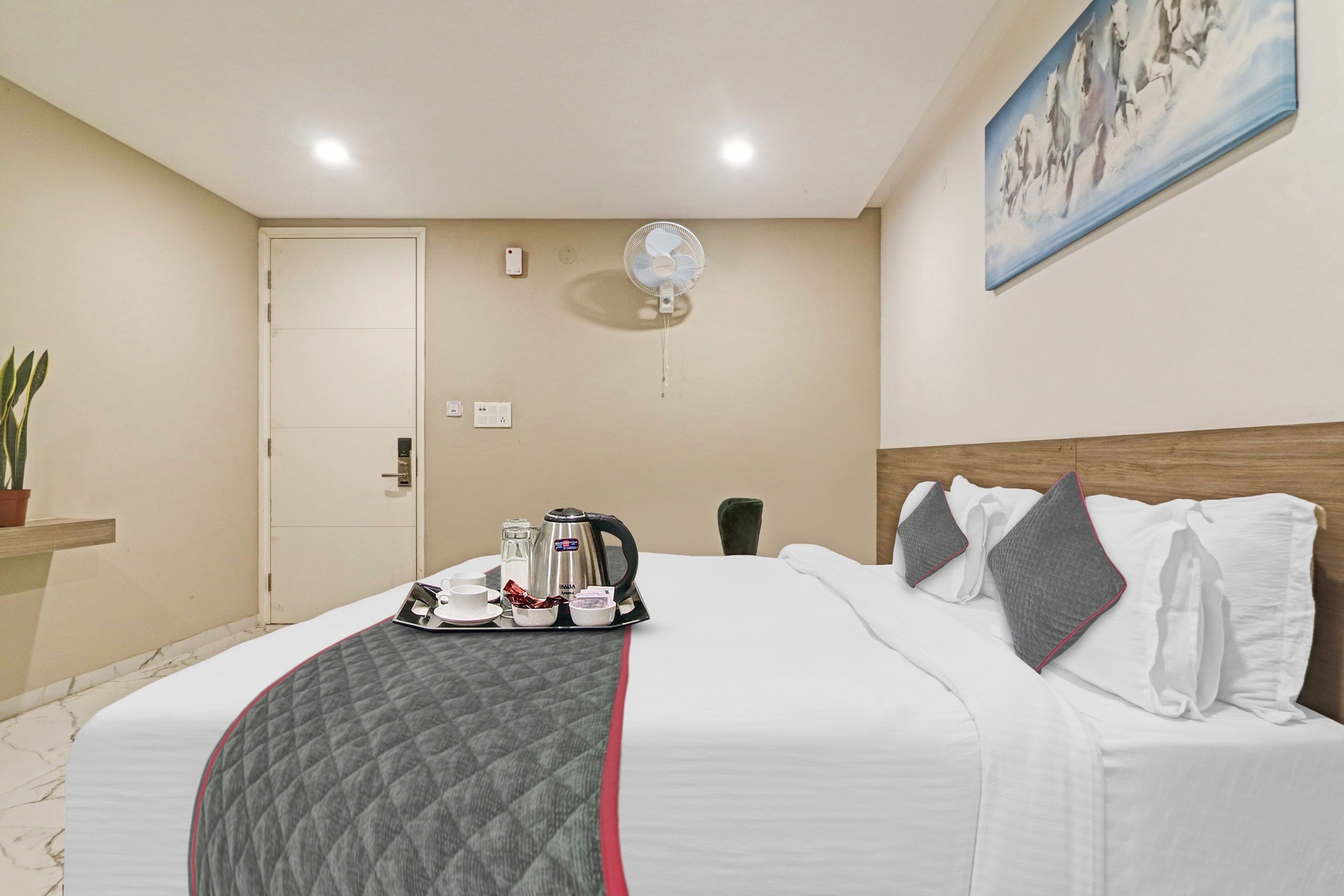 Quarto de hotel com cama, mesa de cabeceira com chaleira e chá, ventoinha na parede e pintura de cavalos.