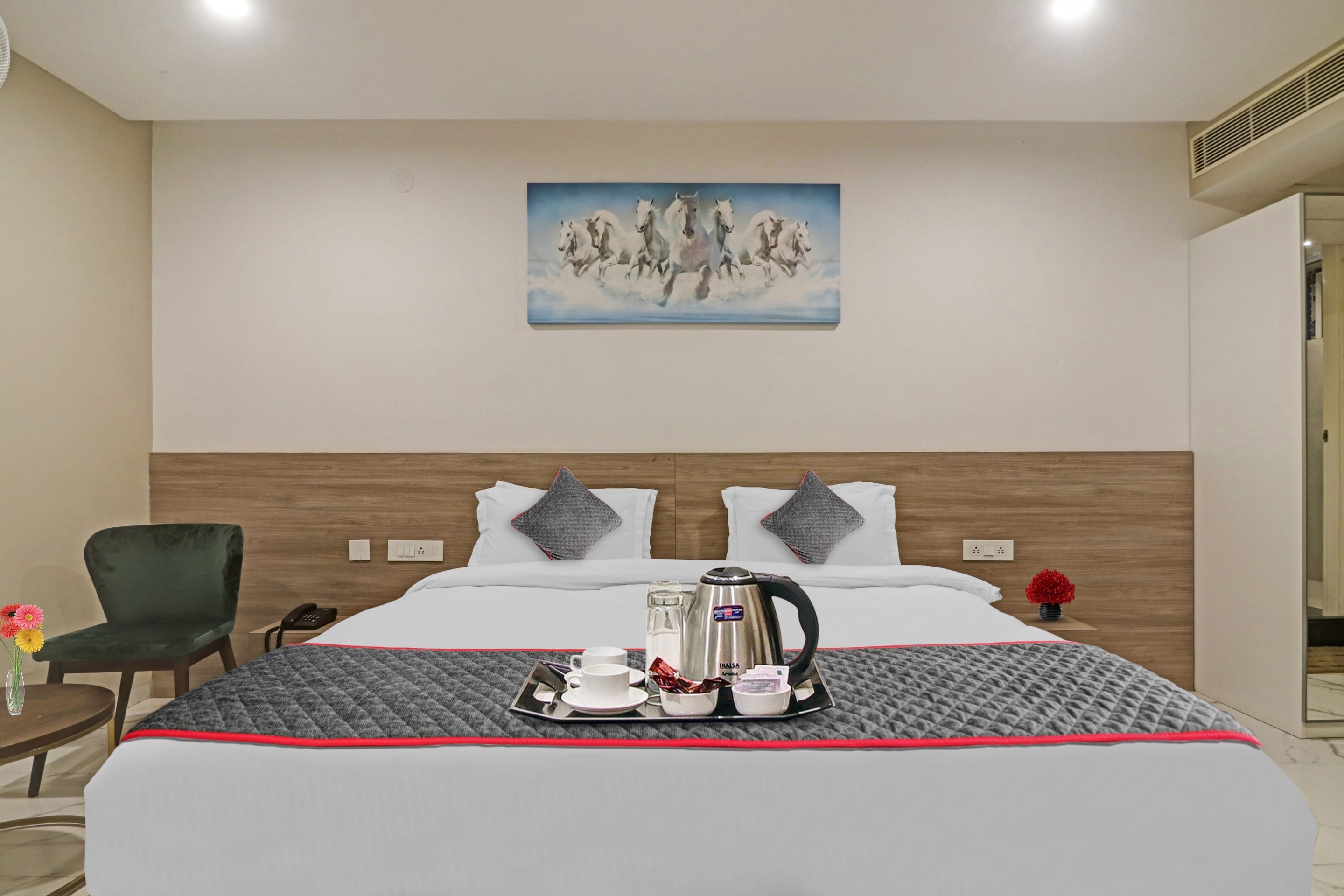 Habitación de hotel con una cama, un cuadro de caballos, una silla, una mesa con una bandeja de desayuno y un jarrón con flores.