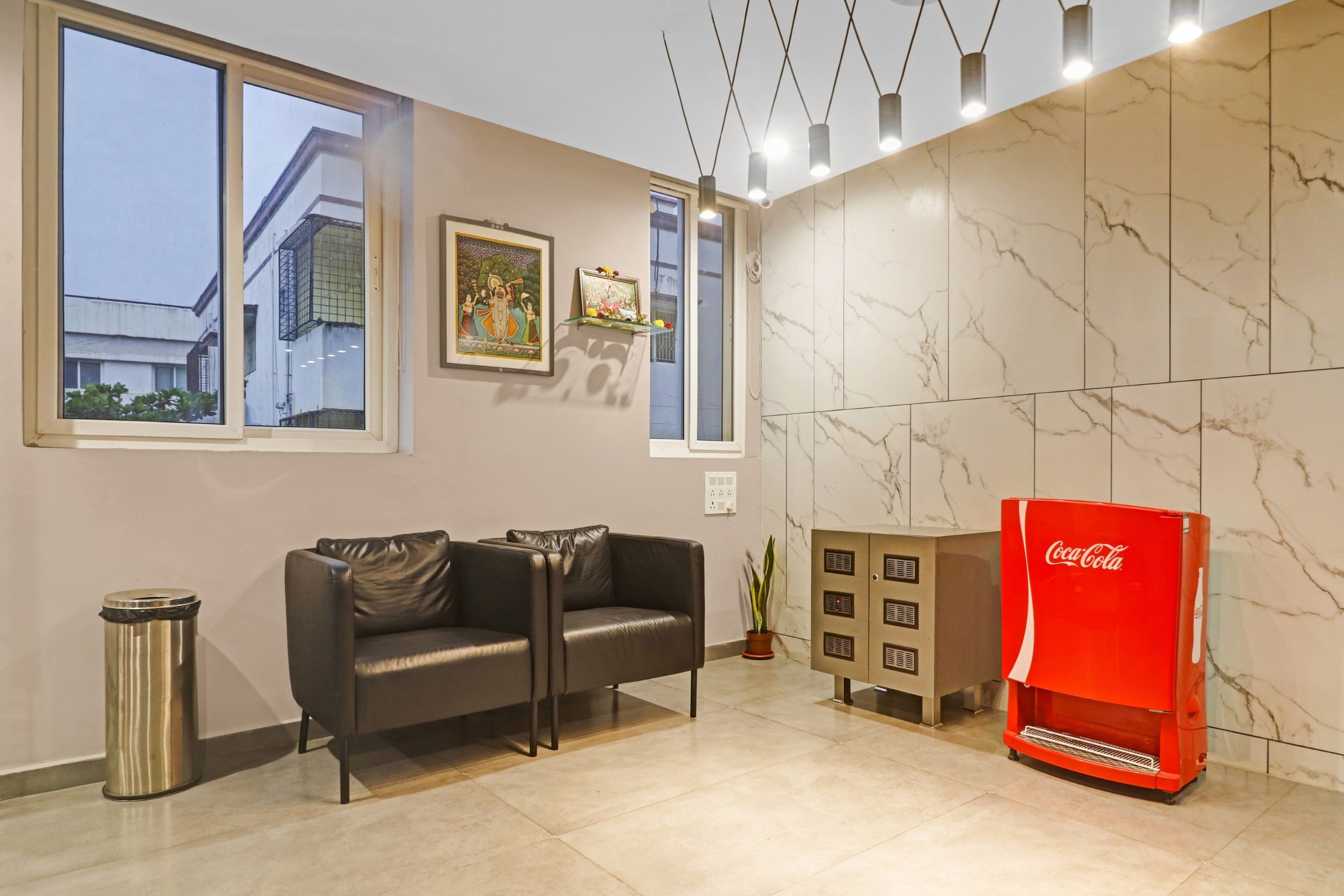 Uma sala de estar moderna com um sofá, uma lixeira, uma geladeira vermelha Coca-Cola e uma janela grande com vista para edifícios.