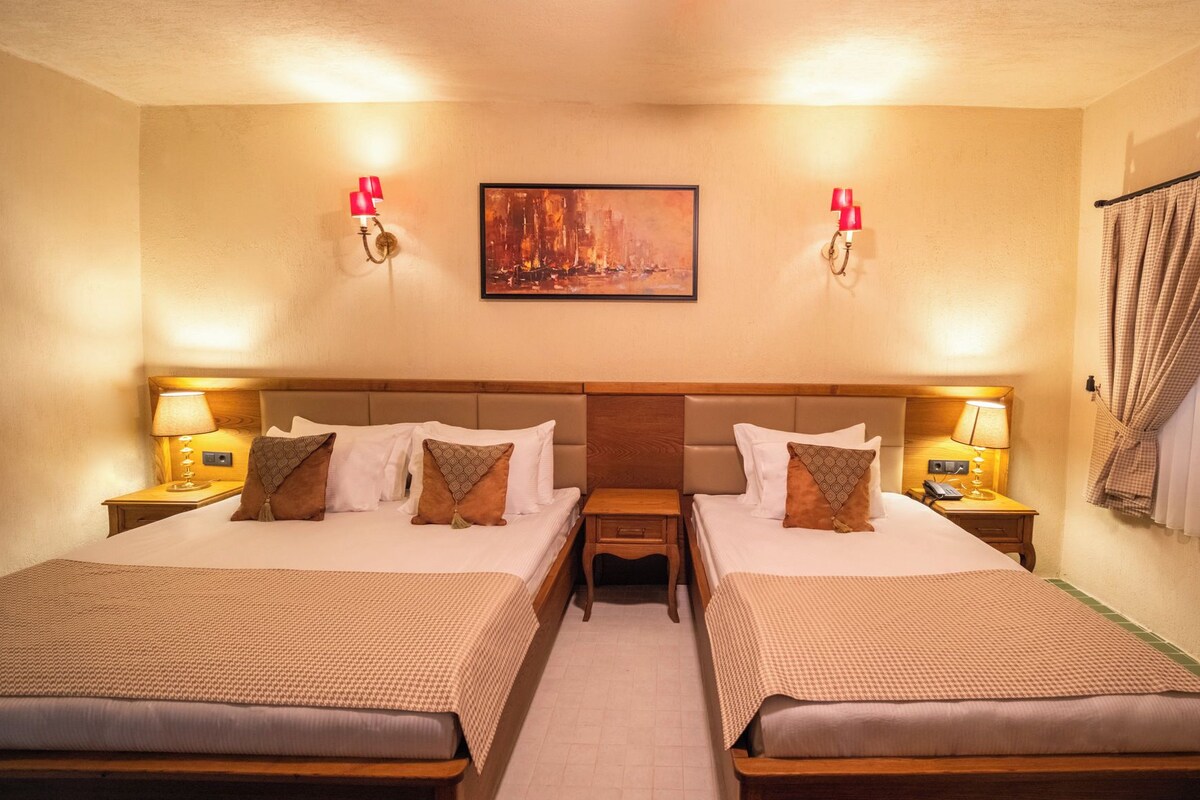 A hotel room with two beds, a painting on the wall, and a window with curtains.