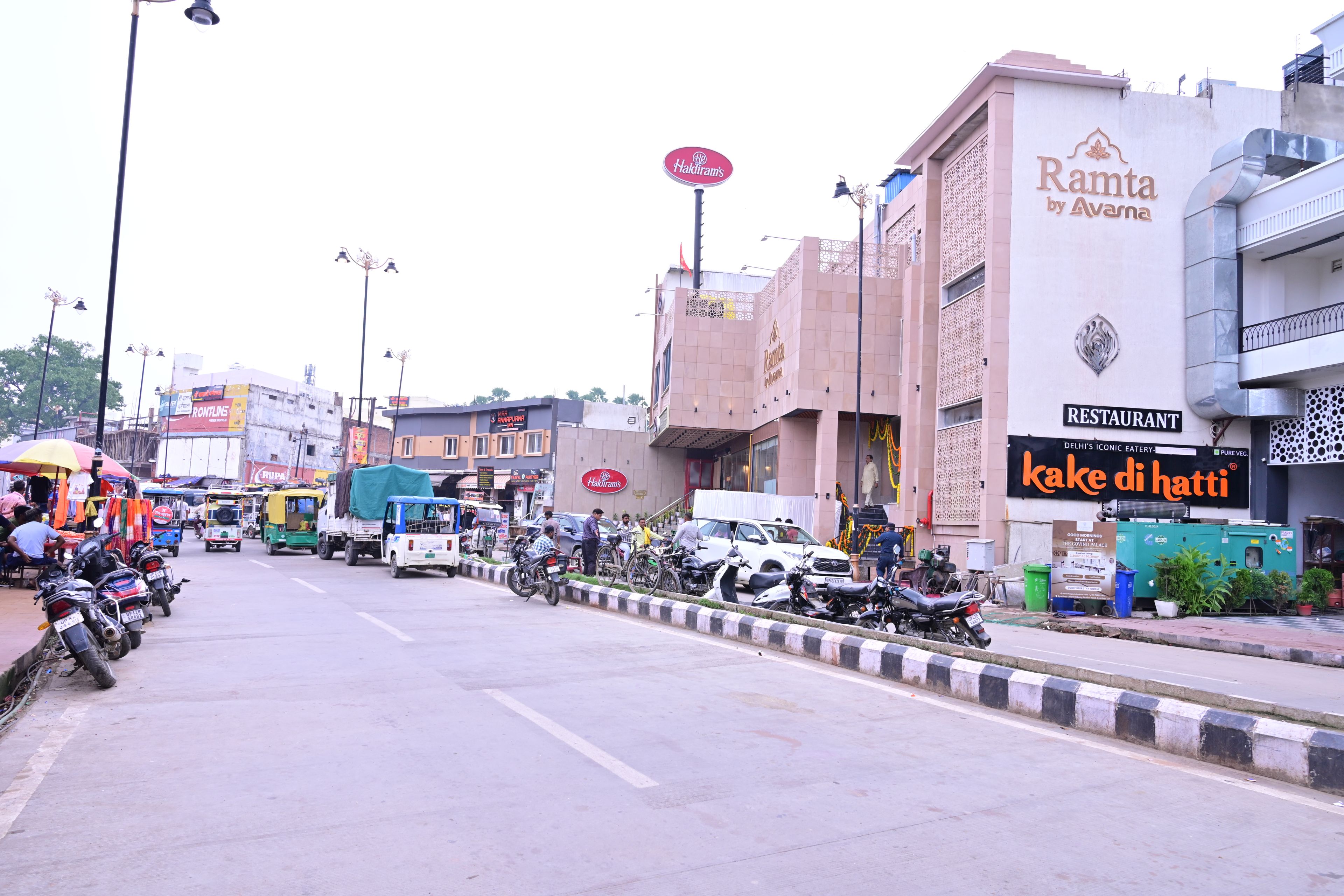 A street with parked motorcycles and vehicles, a restaurant named 'Rama by Avansha' with a signboard, and a building with a sign for 'Haldiram's'.