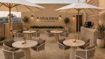 Een terras met ronde tafels en stoelen, parasols en een muur met de tekst 'viva mesa ALL DAY CAFÉ'.