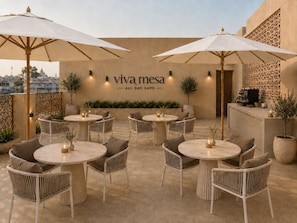 An outdoor seating area with round tables and chairs, umbrellas, and a wall with the text 'viva mesa ALL DAY CAFÉ'. - Hotel Ramta by Avarna (Ayodhya)