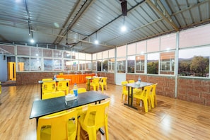 Um espaço amplo e interno com várias mesas e cadeiras, janelas grandes e teto alto.