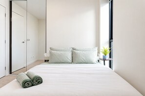 A minimalist hotel room with a large bed, two pillows, a small bedside table with a plant, and a view of the city through a window. - Southbank 2BR 2Bath | 2 Queens & Free Parking (Southbank)