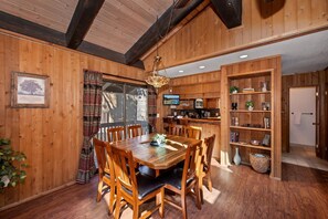 A wooden dining area with a large table and chairs, a chandelier, and a kitchen area with shelves and appliances. - Steps from Snow Summit Resort! Ski In/Out Retreat! Incredible Views of Snow Summit Resort! (Big Bear Lake)