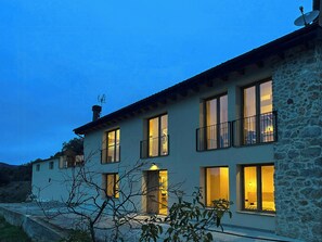 A two-story stone house with multiple windows and balconies, surrounded by vegetation. - Designed pyrenean farmhouse at 1,100 m altitude (Ribera d’Urgellet)