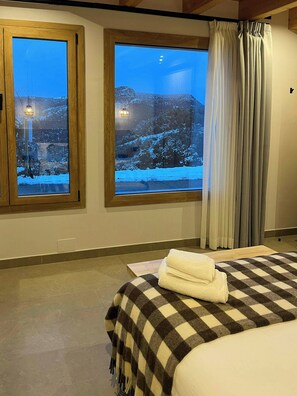 Quarto de hotel com duas janelas amplas, cama com manta xadrez e vista para uma paisagem nevada.