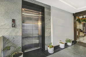 An elevator with a metallic finish, a wall-mounted intercom, and potted plants along the corridor.