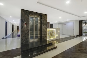 Un ascensor moderno con pantalla digital, un mostrador de recepción con un panel decorativo y un pasillo con puertas y plantas en macetas.