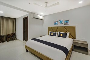 Quarto de hotel com cama, mesa de cabeceira, cadeira, escrivaninha, aparelho de ar condicionado e ventoinha no teto.