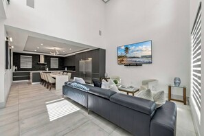 A modern living room with a grey sofa, a dining area, and a kitchen with dark cabinets and a stainless steel refrigerator. - Walk to Raymond James Stadium - Modern Tampa Home with 2 King Beds, Balcony & Outdoor Dining (Tampa)