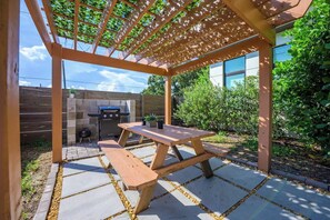 Eine Außenpatio mit Holztisch und -bänken, einem Grill und einer Pergola.