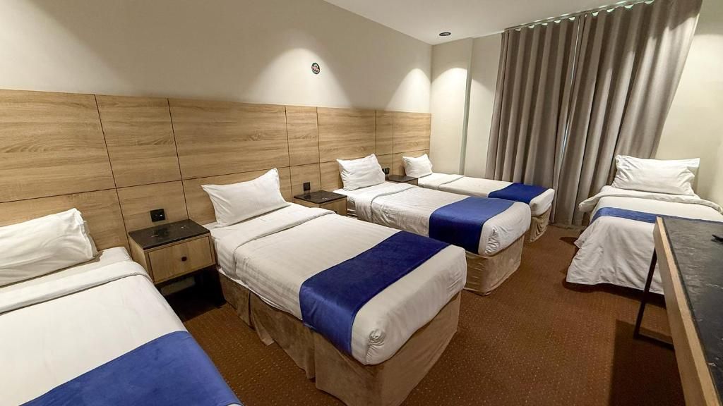 A hotel room with four beds arranged in a row, a wooden headboard, a large window with curtains, and a flat-screen TV on a stand.
