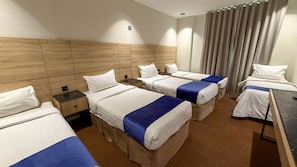 A hotel room with four beds arranged in a row, a wooden headboard, a large window with curtains, and a flat-screen TV on a stand. - Al Massa Al Ayam Hotel (Makkah)
