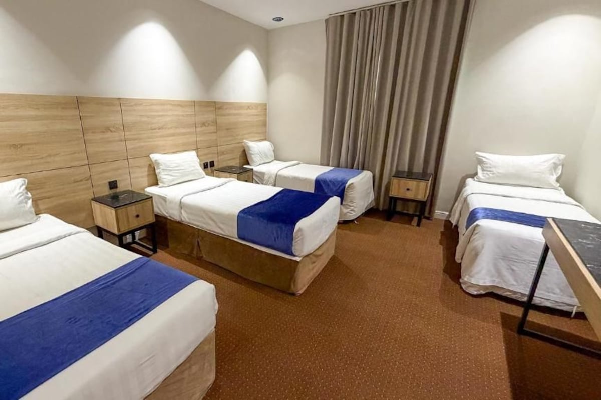 A hotel room with four beds arranged in a row, a wooden headboard, a small desk, and a window with curtains.