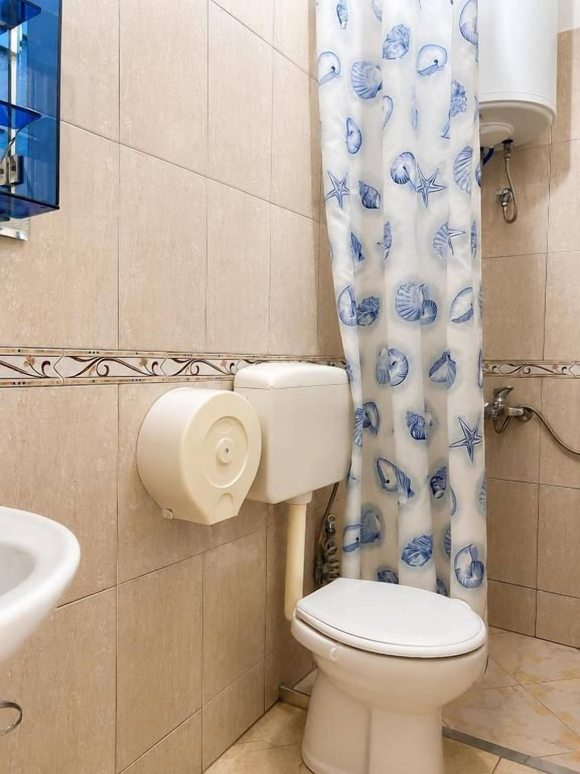 A bathroom with a toilet, a roll of toilet paper dispenser, a shower curtain with a sea shell pattern, and a wall-mounted sink.