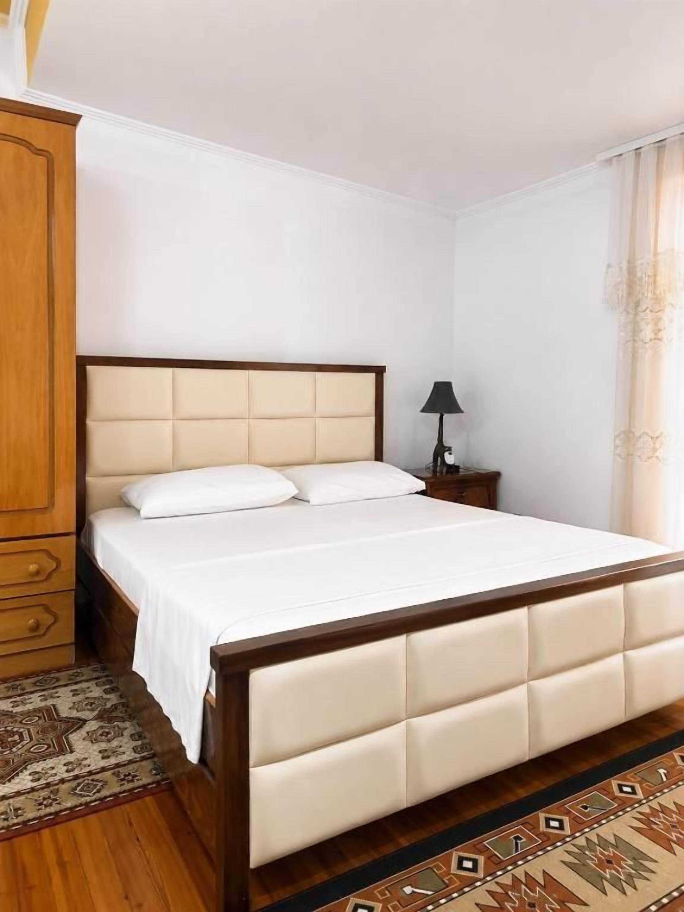 A bed with a wooden frame and headboard, a bedside table with a lamp, and a patterned rug on the floor.