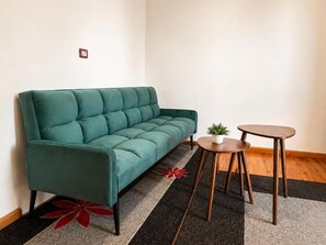 Un sofá color turquesa, una mesa redonda de madera con una planta y una alfombra floral roja en una habitación con suelo de madera y paredes blancas.