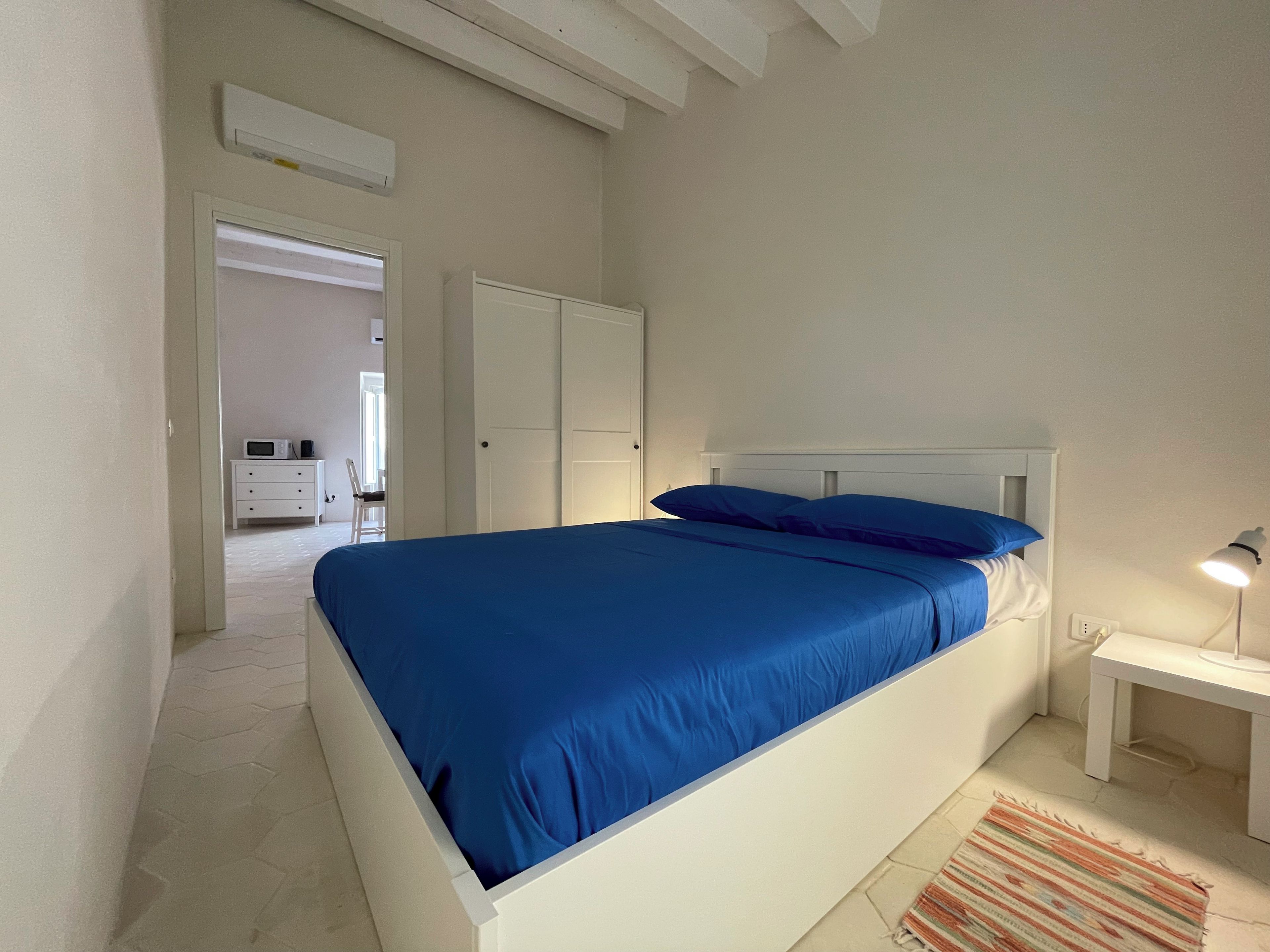 Une chambre à coucher comprenant un lit, une table de chevet, une lampe, une chaise, une armoire et une porte donnant sur une autre pièce.