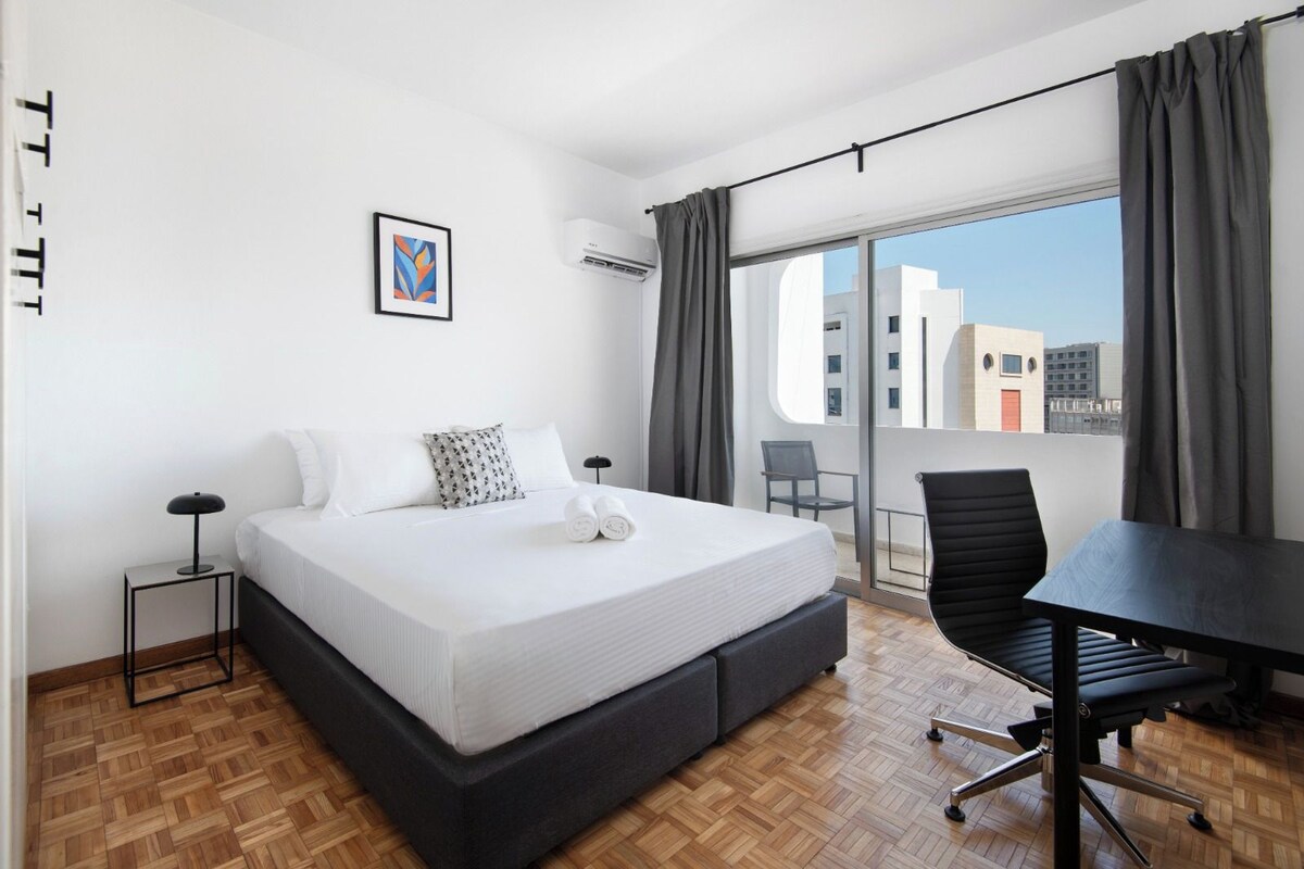 Una habitación de hotel con una cama grande, un escritorio, una silla, una mesita y una ventana con cortinas.
