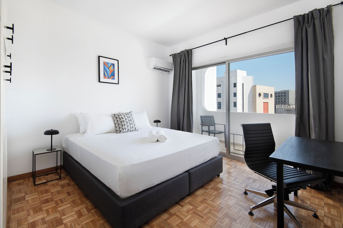 A modern hotel room with a large bed, a desk, and a view of the city.