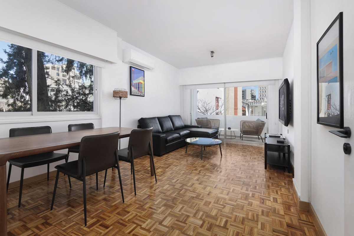 A modern living room with a wooden parquet floor, a black leather sofa, a dining table with chairs, and a television mounted on the wall.