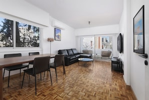 A modern living room with a wooden parquet floor, a black leather sofa, a dining table with chairs, and a television mounted on the wall. - Urban Habitat Executive Suites (Nicosia)