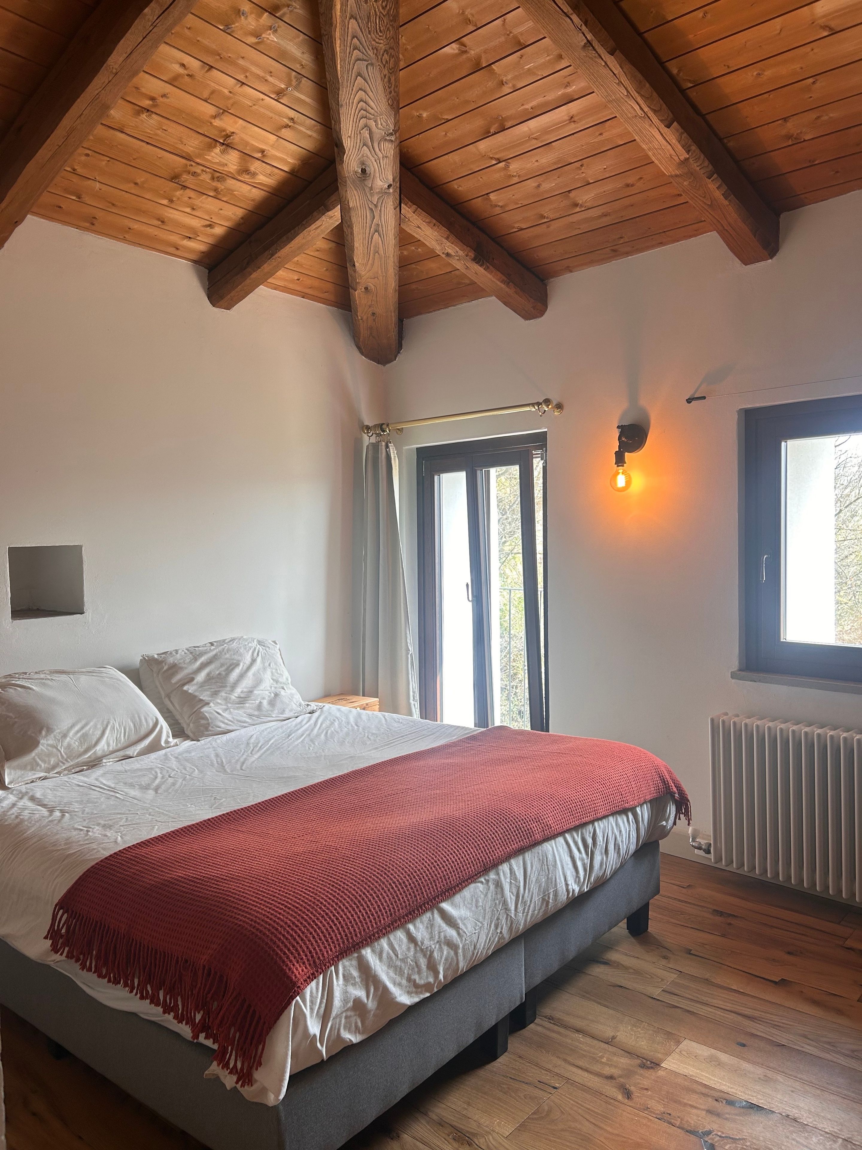 Une chambre avec un plafond en bois, un lit avec une literie blanche et rouge, une fenêtre avec des rideaux et un radiateur.