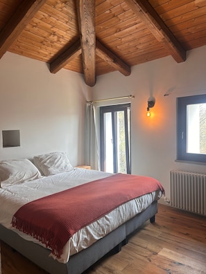 Un dormitorio con techo de madera, una cama con ropa de cama blanca y roja, una ventana con cortinas y un radiador.