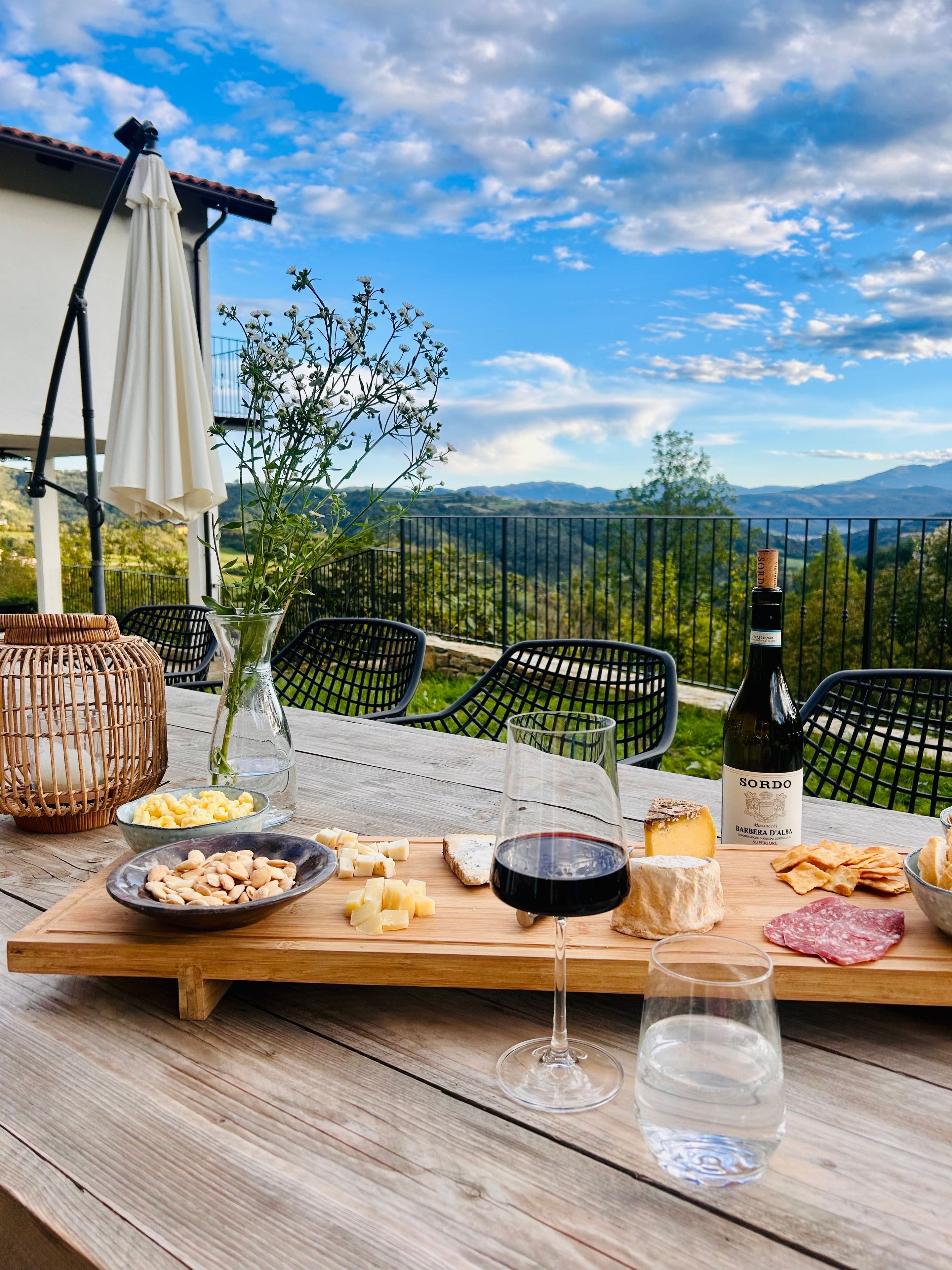 A table set outdoors with a bottle of wine, cheese, nuts, crackers, and a glass of water.
