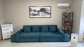 Una sala de estar con un sofá color turquesa, un aparador blanco, un aire acondicionado de pared y un cuadro abstracto enmarcado.