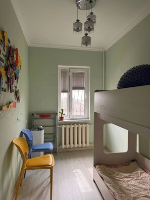 Ein Kinderzimmer mit Etagenbetten, einer Heizung, einem Fenster mit Rollläden und einer Wand mit bunten Verzierungen.