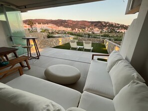 Un espacio de estar moderno al aire libre con un sofá, sillas y una mesa, con vistas a un jardín ajardinado y una vista de un pueblo en la ladera.