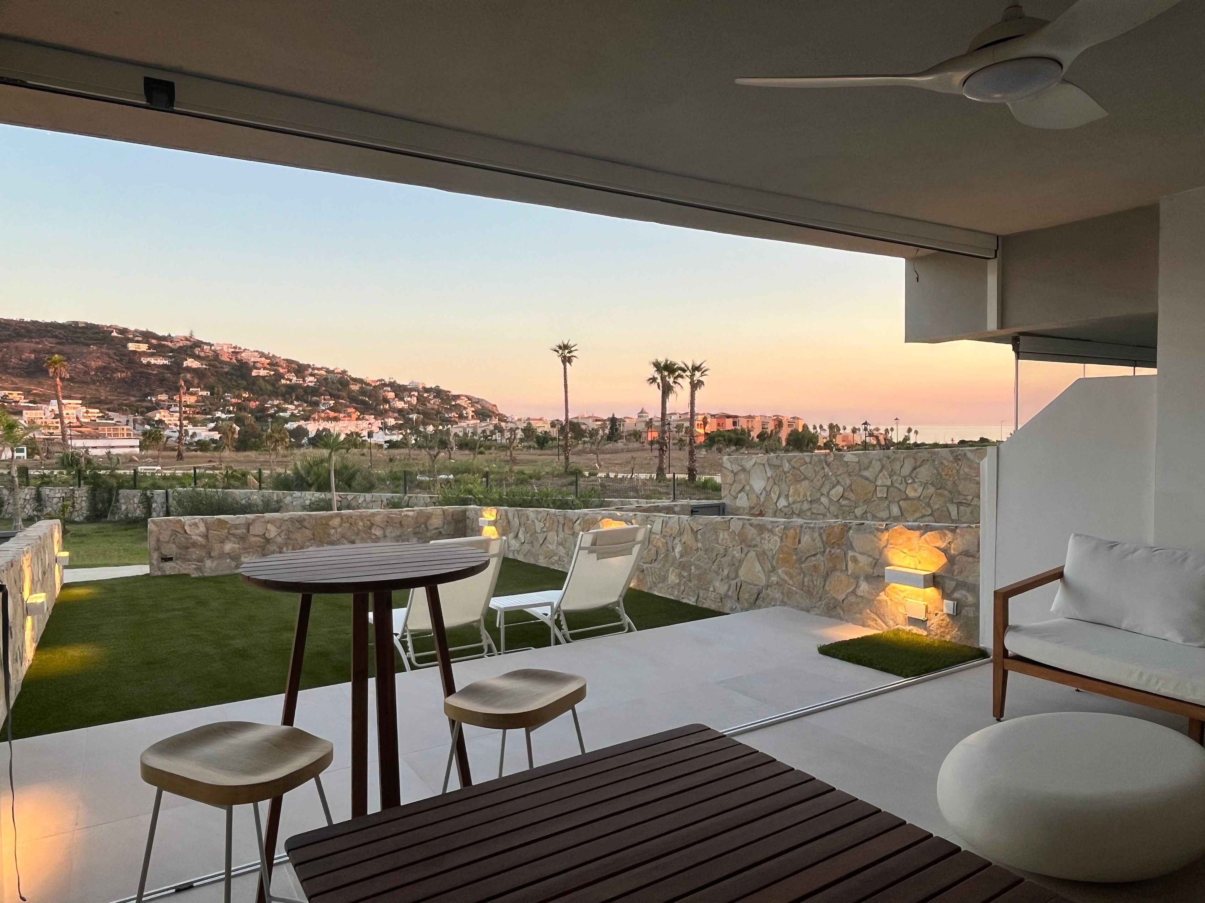 Um pátio moderno ao ar livre com mesa e cadeiras, paredes de pedra e vista para uma vila no topo do morro.