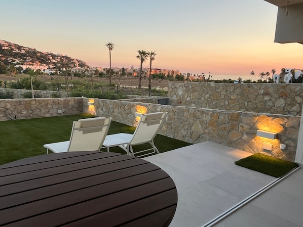 An outdoor patio with a stone wall, two white chairs, a table, and a view of a coastal landscape during sunset.