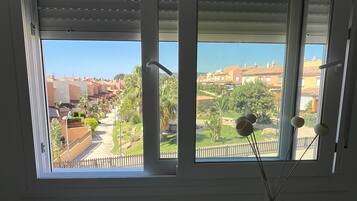 Quarto com escrivaninha branca e cadeira, janela com persianas e vista para uma área residencial.