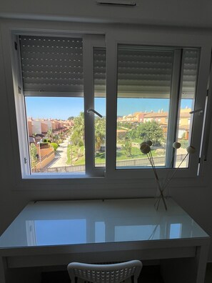 Una stanza con una scrivania e una sedia bianche, una finestra con persiane e vista su un quartiere residenziale.