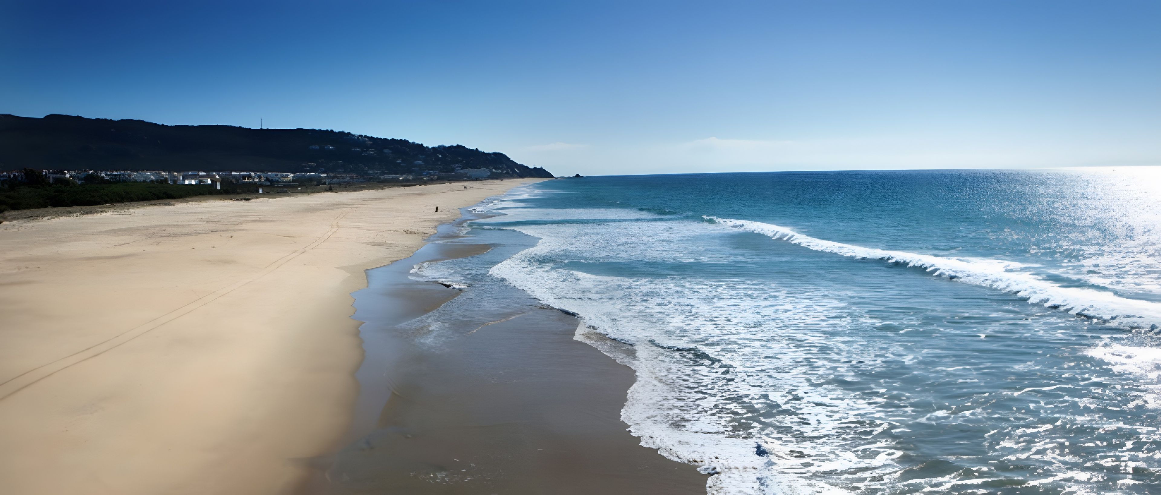 Une plage avec un ciel dégagé, des vagues qui se rapprochent du rivage et une montagne lointaine.