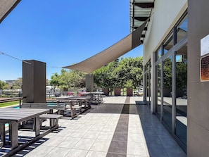 Un espacio al aire libre con patio, mesas, sillas, una piscina y un edificio con grandes ventanales.
