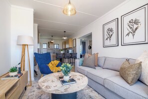 Ein modernes Wohnzimmer mit einer Couch, einem runden Couchtisch, einem blauen Sessel und einer Lampe. Im Hintergrund ist eine Küche zu sehen.
