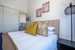 Una cama bien tendida con edredón blanco y almohadas estampadas, una mesita de noche y una lámpara de pared.