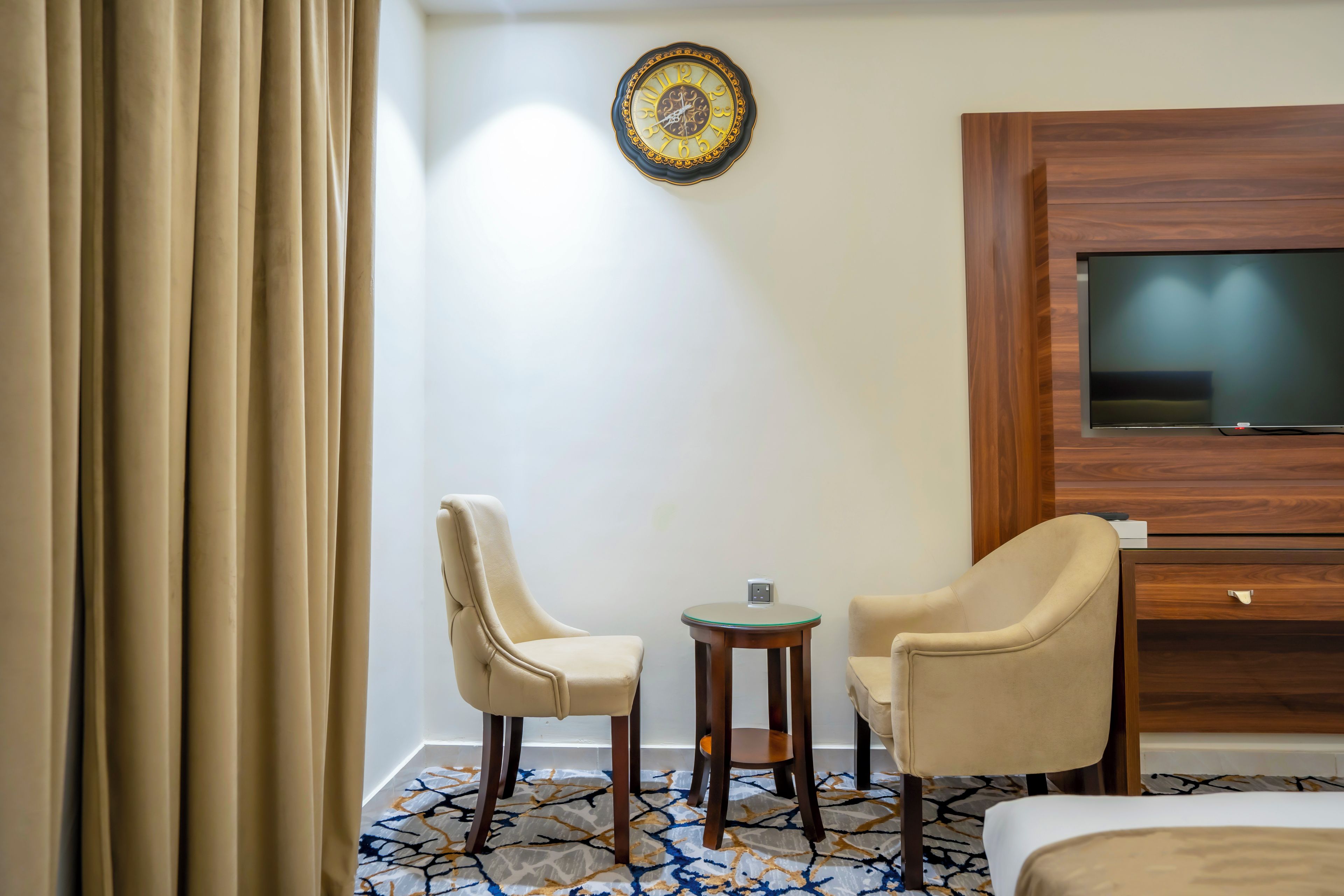 Une chambre d’hôtel comprenant un lit, deux chaises, une petite table, un téléviseur et une horloge murale décorative.