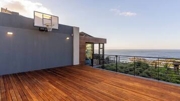 A wooden deck with a basketball hoop, a glass door leading to a covered area, and a view of the ocean.